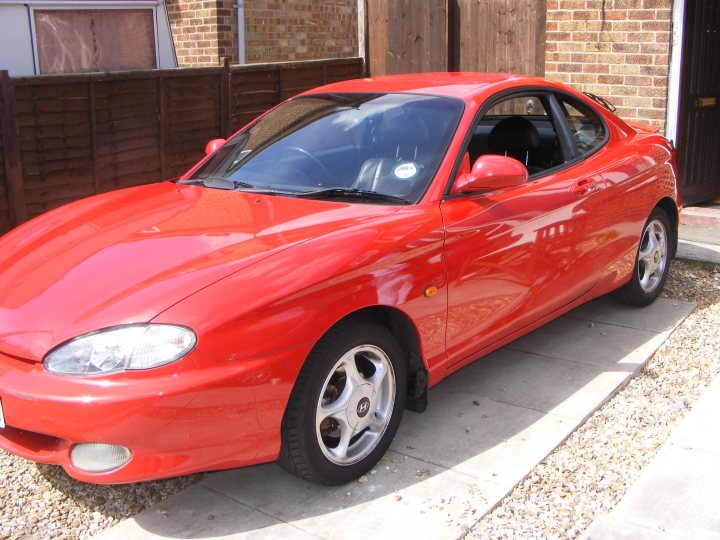 Pistonheads Coupeshed - The image shows a bright red sports or luxury convertible car parked on a sidewalk next to a brick building with a front door. The car is clean and shiny, indicating it is well-maintained. It is a convertible with the top down, revealing the interior seats. The vehicle has alloy wheels, which add to its sleek and polished appearance. The reflection on the car's surface suggests it is a sunny day.