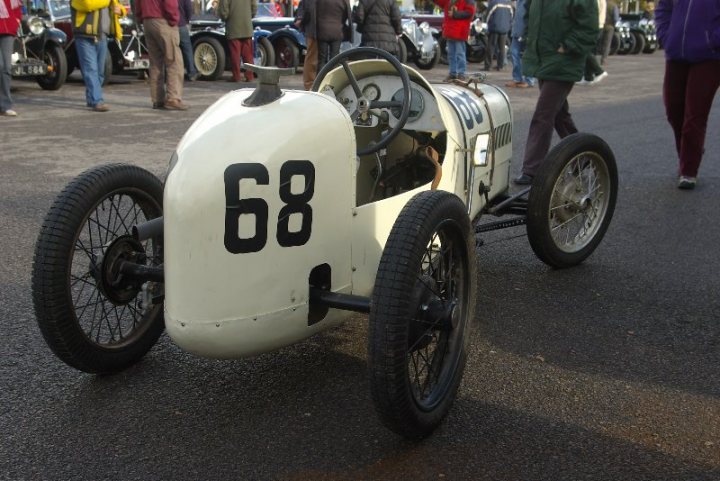 Pistonheads - The image showcases an elegant vintage race car parked on a road. The car is white with distinctive black numbers and details. It's a single seater vehicle featuring two wheels at the front and two at the back. The registration number is clearly visible on the front of the car. The car's white color stands out against the brown of the road and the green and grey hues of the people's attire in the background. The car has a classic racing look, which suggests that it is a rare and valuable specimen.