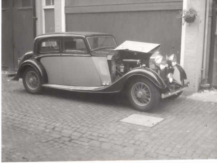 B&W Bentleys.... - Page 1 - Rolls Royce & Bentley - PistonHeads - The image shows an old black and white car parked on a brick road. Its convertible top is down. The car's engine hood is open and the headlights are on, giving it a distinctive look. The background is black and white, hinting at a vintage or historical setting. There is a window to the side, indicating a nearby building.