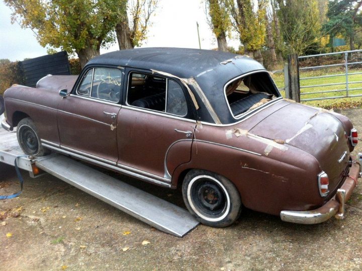 1950's Merc 220s Saved from the grave - rat rod restoration  - Page 1 - Mercedes - PistonHeads - In the image, a classic brown car with a curved front design is prominently displayed. The vehicle is parked on a metal trailer, suggesting movement or storage. A tree stands tall and proud in the background, adding a touch of nature to the scene. The overall setting appears to be outdoors in the daylight. The car has a rusted roof and a tailgate, indicating possible signs of aging or exposure to the elements. The rustic charm and vintage aesthetics of the car contrast with the stark, angular design of the trailer and the rigid, vertical trunk of the tree.