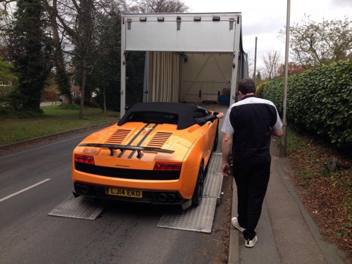 Performante Edizone Tecnica  - Page 1 - Gallardo/Huracan - PistonHeads - In this image, a man is seen walking alongside an orange sports car on a street. The car is being loaded onto a trailer by a car transporter truck. The carrier truck appears to be in motion on the street, with an opening in the back, revealing an interior. In the background, there's a beautiful landscape with a lush greenery of trees and bushes. The man's attire suggests he might be the owner or handler of the car.