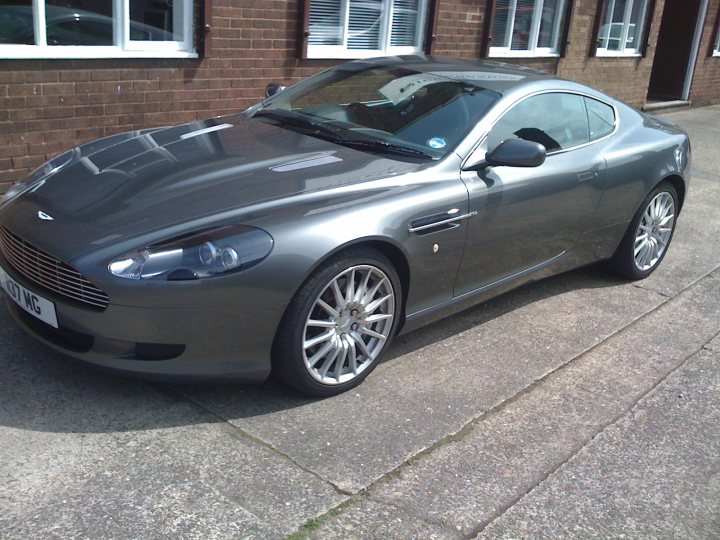 Sub 20k DB7 - Page 1 - Aston Martin - PistonHeads - The image depicts a silver Aston Martin sports car parked on a sidewalk outside what appears to be a residential building. The car is sleek and stylish, with the distinctive Aston Martin logo prominently displayed on the grille. The background is mainly composed of the brick wall of the building, which sets a contrasting tone to the shiny metallic finish of the car. There are no visible people or animals in the image, and the focus is solely on the vehicle.