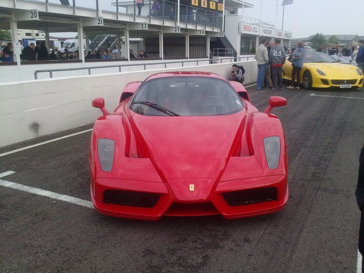 Pistonheads Supercar Sunday - This image captures a vibrant scene on a race track. The most prominent feature is a sleek, red Ferrari sports car, which takes up the center of the frame. Its streamlined design and bright color suggest it's designed for speed and performance. The tracks are lined with spectators, some of whom are positioned near what appears to be a pit or station area, while others are scattered around the track area, adding to the atmosphere of the event. The backdrop of the dilapidated building juxtaposes the modern sportscar, hinting at the contrast between heritage and innovation in the automotive world.