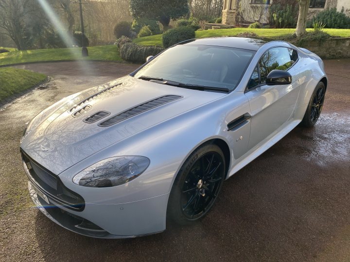 The non-black-non-grey-non-silver thread - Page 48 - Aston Martin - PistonHeads UK - The image displays a silver sports car parked on what appears to be a wet road. The car is sleek and modern, with a prominent front grille and large wheels. It has the distinctive design elements of an Aston Martin vehicle, such as the side gills and the low and wide stance typical of high-performance vehicles. The setting suggests it might have rained recently, as the road surface looks wet. In the background, there is a grassy area with some foliage visible. There are no people in the image, and the car is the central focus.