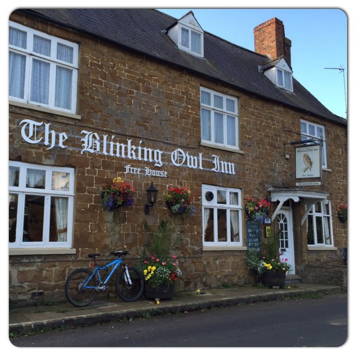 The "Photos From Today's Ride" thread... - Page 225 - Pedal Powered - PistonHeads - The image depicts a quaint, thatch-roofed pub called "The Blind Owl Inn". The signboard above the entrance is white, adorned with the inn's name in elegant, dark cursive. Flanking the entrance are multi-colored flower boxes, adding a touch of vibrancy to the otherwise stone facade. A blue bicycle is parked in the foreground of the image, resting against the wall of the inn. The scene suggests a charming, inviting atmosphere, possibly located in a countryside setting.