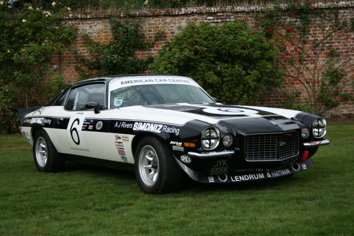 Minilite wheels love -in. - Page 11 - Classic Cars and Yesterday's Heroes - PistonHeads - The image shows a classic muscle car with a black and white color scheme. It's parked outdoors on grass, suggesting an event or show. The car is adorned with various decals, including sponsor logos and numbers, which indicates it may be involved in a racing or competition context. The setting includes a brick wall and a tree in the background, giving the impression of a park or a similar outdoor space. The overall style of the image is a standard photograph with no visible filters or artistic manipulations.