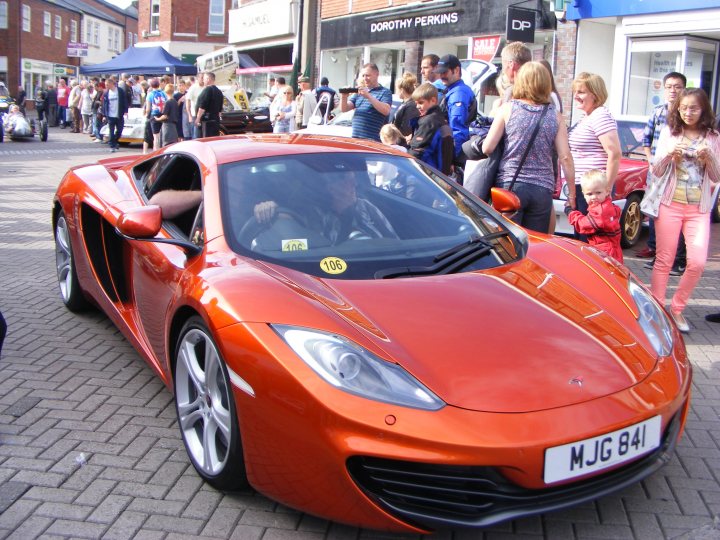 Ormskirk Motorfest, Sunday 26th August. - Page 1 - North West - PistonHeads - The image captures a vibrant scene from a city street where a luxury sports car, painted in a striking shade of orange, is parked amidst a crowd. The car's sleek design and shiny finish are prominently visible, making it the focal point of the image. Several people are gathered around the car, some closer than others, their attention captured by its allure. They are casually dressed, with a few carrying handbags, suggesting a relaxed, social atmosphere. The backdrop of the image reveals a typical urban setting with other cars and buildings, indicating that this is a street in a town or city.