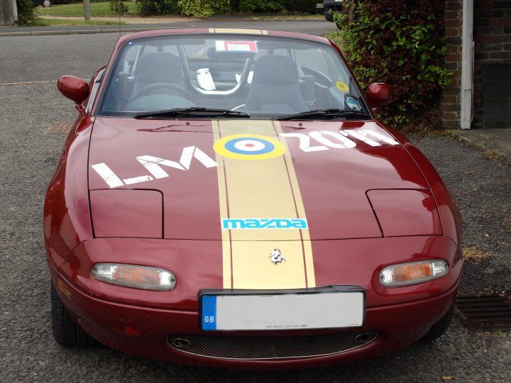 Stickering up for 2016 - Page 1 - Le Mans - PistonHeads - The image shows a red Mazda car parked on a driveway. The car bears a sign that reads "LMBC 201", signifying its involvement in the London Marathon clock collection program in the year 2011. Additionally, there is a large decal on the car's hood depicting the London Marathon logo and the text "MIRACLES MILLIONS 2011". The car appears to be in good condition, suggesting it's well-maintained or even possibly being used for promotional purposes. The setting is an outdoor residential area, with the driveway visible and a portion of some vegetation in the background.