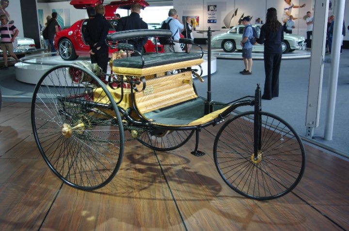 Anyone been to the Moving Motorshow before? - Page 1 - Goodwood Events - PistonHeads - This image showcases a historic quad limousine on display in a museum exhibit. The vehicle, painted in a combination of red and yellow, is an early example of automotive transportation. Its design includes four oversized wheels and a yellow back seat area. Spectators can be seen touring around the vehicle, admiring its classic design and features. In the background, additional vintage cars and booths contribute to the educational atmosphere of the museum.