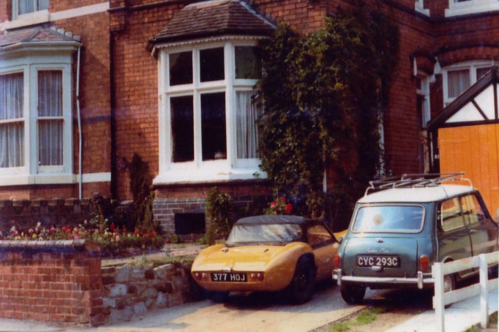 A car parked on the side of the road - Pistonheads - The image captures a peaceful street scene with several vintage cars parked along the curb. The dominant colors are warm, with reds, yellows, and browns, indicative of a classic or antique setting. The architectural style of the building, with its brick facade and white-framed windows, adds to the nostalgic ambiance. Two distinctive cars are parked in front of the building: a small yellow car and a classic green car that has a British license plate. The setting seems to evoke a sense of timeless charm and simplicity.