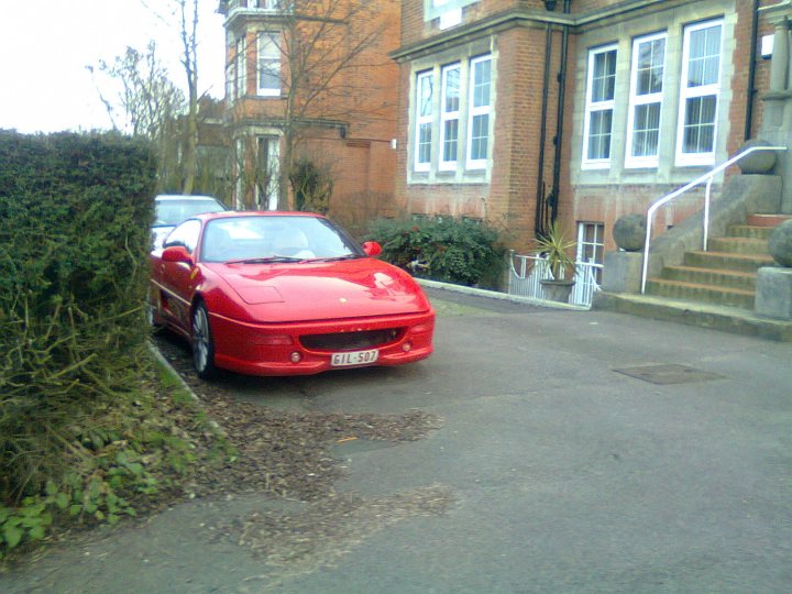 Is this fake? - Page 1 - General Gassing - PistonHeads - The image depicts a residential street scene. A red sports car is parked on the driveway of a house with a tall, narrow front door on the top right corner. The car has a yellow license plate. There is a well-maintained green hedge to the left of the car, partially obscuring the view of its front. In the foreground, there is a narrow, paved street lined with small bushes and shrubs. The sky appears overcast with a white, washed-out look, suggesting either dawn, dusk, or a recent rainfall.