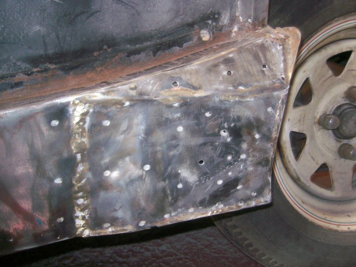 Pistonheads - The image shows a close-up view of a vehicle's suspension system. A damaged metal part is sandwiched between the tire and the vehicle's body, with several bolt holes visible. Rust stains and protruding metal edges suggest wear and corrosion. The focus is on the metallic components, with the tire partially visible in the background.