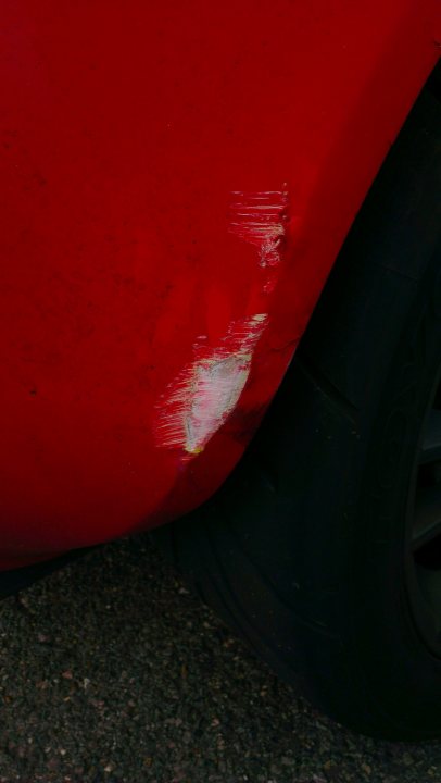 Bugger! Hit a wall - damaged rear clam - advice? - Page 1 - Elise/Exige/Europa/340R - PistonHeads - The image features a close-up of the rear left wheel of a red car. At the top of the wheel, there is a noticeable scrape with chips of red paint. In the lower right corner, there is a reflection on the black asphalt, showing the visible part of the wheel and a portion of the car's body. The focus of the image is on the automobile's damage and the detailed texture of the tire's rubber.