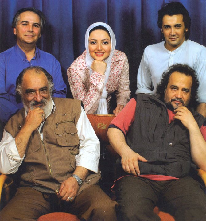 In this image, five individuals are seen posing together against a blue background. A man is seated on a chair on the left side of the frame, smiling at the camera. Next to him, to his left, a woman is seated, donning a hijab and a traditional dress, pointing at something in the room. Beside her, another man is seated, looking directly at the camera with a side-smile. To the right, two more men are visible; one is seated near the center of the image, also smiling, and the last one is standing towards the right, also posing for the camera. The overall atmosphere seems casual and relaxed, with no text present in the image.