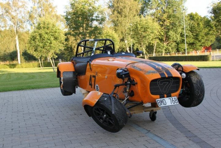 caterham racing colour code? - Page 1 - Caterham - PistonHeads - The image showcases an orange four-wheeler ridden by a single individual. The person is wearing a yellow vest for safety. The ride is equipped with large black tires and is parked on a brick road. In the background, there are trees and a fence, indicating a public outdoor space. The perspective of the image is from the driver's side, giving a clear view of the vehicle and its surroundings.