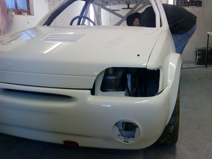 Pistonheads Saloon Northern - The image shows a white car that appears to be undergoing restoration or modification. The front part of the car is prominently visible, with the focus being on the repair area. There's a person wearing a black jacket crouched over the hood, seemingly examining or working on the vehicle. The word "Drink" is written on the surface of the car, suggesting that it might be in a drink drive testing process or is related to some promotional activity. The scene takes place inside a garage or workshop setting. The lighting in the photo is bright, casting a professional and industrial feel to the composition.