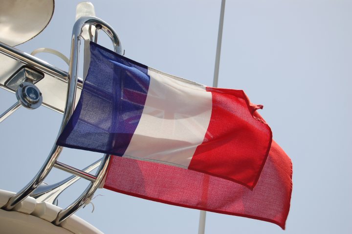 Feel French Pistonheads - The image showcases the French flag. The flag's vertical stripe of blue stretches out, with the white vertical stripe running parallel, followed by a significant arc of red near the folding point of the flag. Adjacent to the flag, there's a silver sailing pole with a white top, installed on a white boat, perhaps hinting at a nautical setting. The flag waves proudly against the sky, indicating its prominence on the vessel it's mounted on.