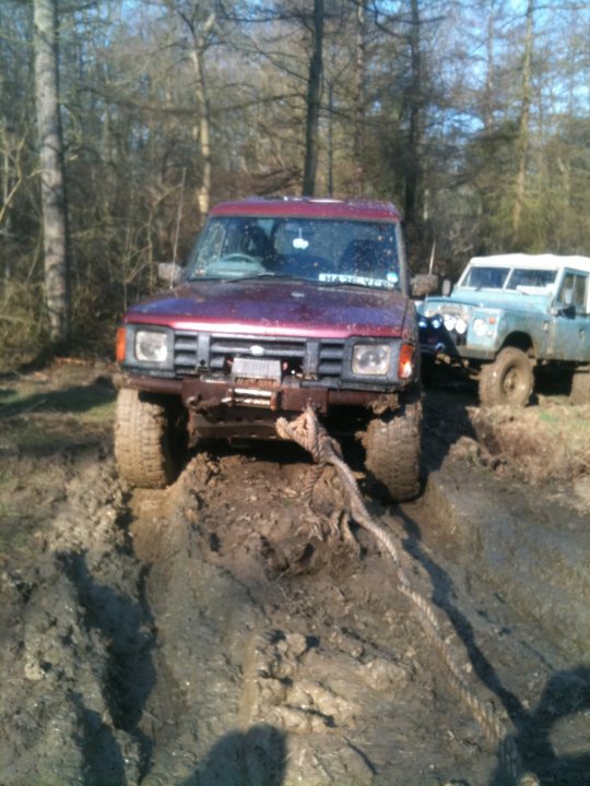 Pistonheads - The image shows a red off-road vehicle partially submerged in mud, with a large log laying over its hood and mud flaps. In the background, another off-road vehicle and trees indicate a woodland setting. There is no visible text or branding in the image. The terrain suggests that the vehicles are in a muddy area, likely meant for off-roading. The vehicles appear to be in use, possibly participating in or preparing for a mud run or similar activity.