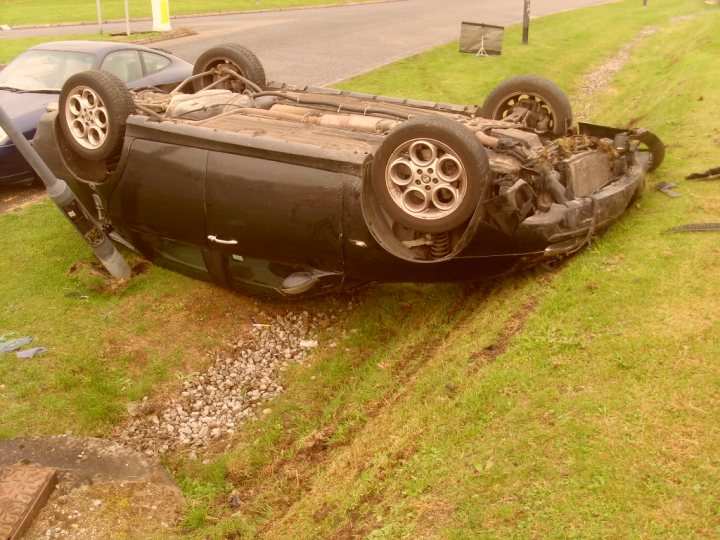 Rover and out! Cribbs crash. - Page 1 - South West - PistonHeads - The image depicts a black car that has been severely tipped over and landed upright in a grassy area. The car's roof appears to have ripped away, revealing the interior. The scene suggests the aftermath of a car accident, with the vehicle's roof missing and the damage indicating a significant impact. The car is stationary, possibly left in the same position after the accident. The grass around the car is green, and there is no immediate sign of intervention or further damage.