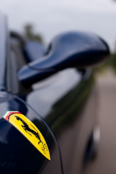 What to Buy -F355-360-F430??? - Page 6 - Supercar General - PistonHeads - The image shows a close-up of a car's side mirror, taken from a slightly elevated angle. The side mirror is black, and it is situated near the rear of the vehicle, angled outward. The background of the image is partly cloudy and mostly blurred, but it seems to be taken on a road or surface appropriate for vehicles. There is a license plate on the back of the car, but it has not been clearly visible in the image. At the bottom of the mirror's frame is a red Ferrari logo, adding a splash of color to the otherwise monochromatic scene.