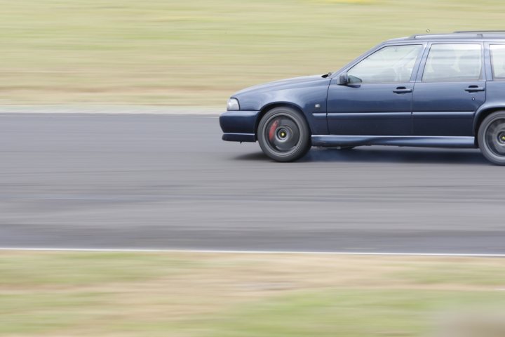 Tyre West Fitters Pistonheads Intro Newbee Suffolk - In the image, there is a black sedan speeding down a curved road. The car appears to be in motion, moving from the left side of the road to the right, and the background is blurred due to its high speed. The road is paved and seems to be in a rural or suburban area, as indicated by the grassy verges on either side. The image is slightly blurry, adding to the sense of the car's speed and the road's curvature.