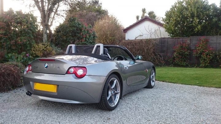 BMW Z4 3.0 auto 2005 or earlier. What are your thoughts?  - Page 2 - BMW General - PistonHeads - The image depicts a metallic grey convertible sports car parked on a gravel driveway. The car has a sleek design with black accents and a black fabric roof that is folded down, indicating it's a convertible model. The vehicle is positioned facing the camera with the front of the car clearly visible, and the top half of its body can be seen.

The driveway is bordered by a wooden fence and lush green shrubs, suggesting a suburban or residential setting. In the background, there is a small white house with a pitched roof, partially obscured by vegetation. The driveway is clear, leading up to the house, with no visible parked cars or people. The sky is overcast, casting a soft light on the scene, enhancing the metallic sheen of the car.