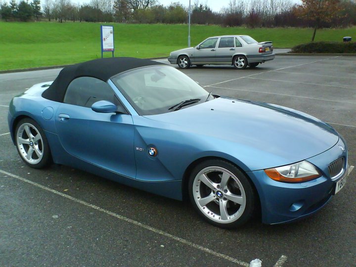 BMW Z4 - Page 1 - Readers' Cars - PistonHeads - The image shows a striking blue BMW Z3 roadster parked in a parking lot during the day. The car has a convertible top down, giving it a sleek, aerodynamic look. It is positioned against a backdrop of a field, and there is a blue sign in the parking lot, possibly a parking direction sign. The sky overhead is overcast, and the ground is paved. Another vehicle with a black roof is partially visible in the background, suggesting the presence of other cars in the lot.