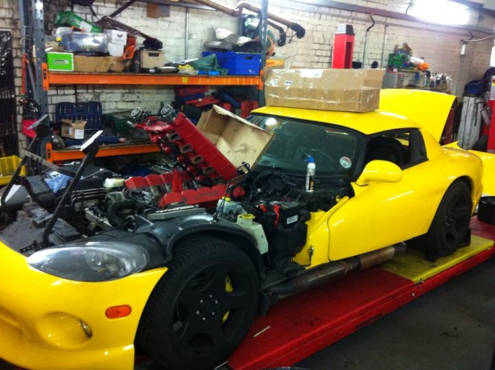 A red motorcycle is parked in a garage - Pistonheads - The image depicts a bright yellow Porsche GT4 lying on its side atop a red and yellow stainless steel frame. Its engine is out, exposing the parts. The car is in a workshop with various tools and equipment scattered around in the background.