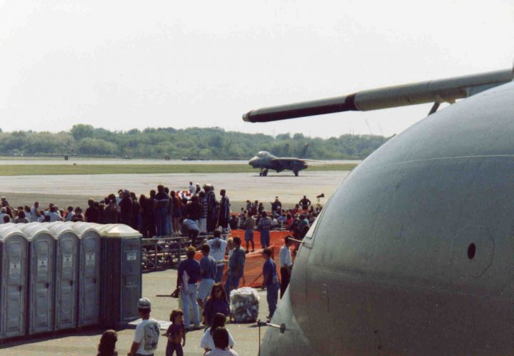 Post amazingly cool pictures FROM aircraft. - Page 17 - Boats, Planes & Trains - PistonHeads - The image captures a lively scene at an airfield. A large military aircraft is parked on the tarmac, dwarfing the crowd of onlookers that surround it. Numerous large portable toilets are arranged haphazardly, suggesting a significant event or gathering is taking place. A fence with racer flags is also visible, adding to the festive and high-energy atmosphere. The background provides a lush expanse of trees, contrasting with the man-made structures and aircraft.