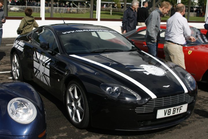 Stickered Pistonheads - The image captures a scene at a car show with a striking black and white Aston Martin DB9 as the main focus. The DB9 is decorated with British flags and other decals on its side and hood. In the background, there are several other cars, including a vibrant red sports car and another one with a white sports car. People are gathered around the cars, with two men visibly admiring the Aston Martin vehicle. The cars are parked on a track, and there are spectators who appear to be enjoying the event.