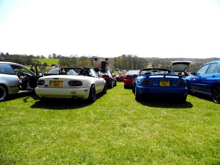 BHP Show - Who's going? - Page 1 - Kent & Essex - PistonHeads - The image captures a lush green field where several cars are parked side by side, bathed in the warmth of a sunny day. The cars range in color from pristine white to vibrant red and deep blue. The open trunk of a white car and the convertible top of a blue car suggest that these vehicles are ready for adventure. A few people can be seen in the background, adding a human touch to this scene of automotive beauty. The emerald expanse of the field and the sky, dotted with clouds, provide a serene backdrop to this gathering of automotive enthusiasts.