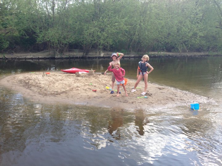 Local lake or river for swimming? - Page 1 - Thames Valley & Surrey - PistonHeads - The image shows three children on a sandy riverbank, which appears to be on horse shoes. There are beach toys scattered around, indicating they might be playing. A canoe is visible in the background, and the water is calm. The children are in casual clothing, with two of them appearing to have their arms crossed. The forest is in the background, suggesting a tranquil natural location.