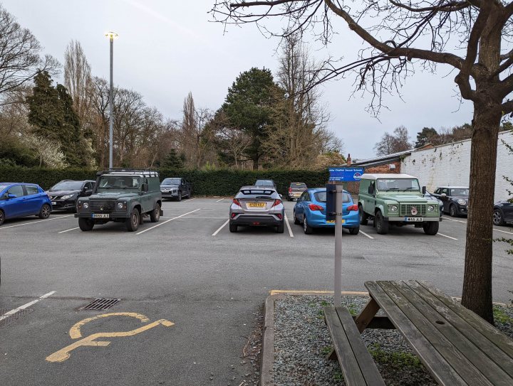 Real Good Number Plates Vol. 6 - Page 325 - General Gassing - PistonHeads UK - The image depicts an outdoor parking lot at a facility with a blue sign, suggesting it may be a hospital or healthcare-related building. There are several cars parked in the lot, and a bench is visible to one side. In front of the building, there's a sidewalk where pedestrians can walk, and beyond that, a tree can be seen. The sky appears overcast with no visible people.