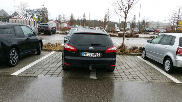 The BAD PARKING thread [vol3] - Page 449 - General Gassing - PistonHeads - This image captures a scene in a parking lot on what looks like a rainy day. Several cars are parked diagonally along white lines, but there's a black car parked sideways, seemingly too large to fit in a standard parking spot. Leafless trees without their foliage can be seen scattered throughout the lot, possibly suggesting autumn or winter, and adding to the slightly moody atmosphere. A building with red, white, and blue stripes can be glimpsed in the background, blending a bit of color into the scene.