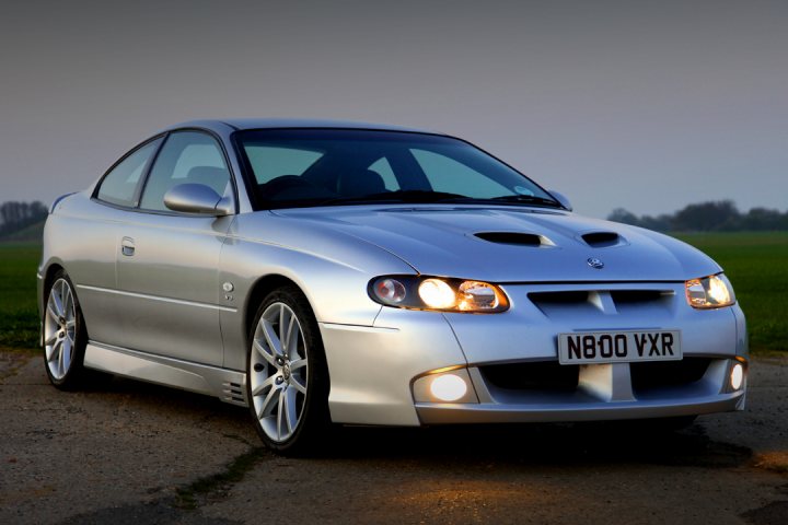 Best picture of your car - Page 9 - HSV & Monaro - PistonHeads - The image features a sleek silver sports car parked on a road during dusk. The car has a streamlined design with modestly sized wheels and a visible grille, which is illuminated at the front. High beams add a distinct ambiance to the scene. The background shows a grassy area under a gray, overcast sky, suggesting a calm and peaceful environment.