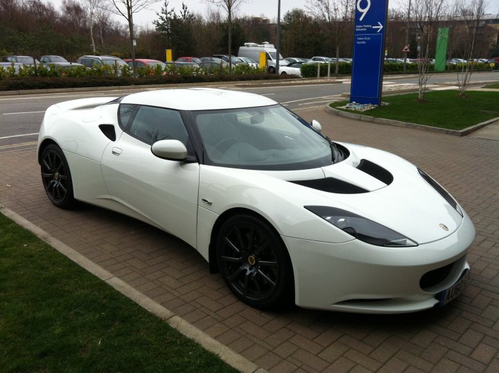 Random Evora Pr0n... - Page 1 - Evora - PistonHeads - The image shows a stylish, white sports car parked outdoors on a gray, tarmac surface. The car has a sleek and aerodynamic design, with a prominent hood bulge, black alloy wheels, and a low roofline that rises slightly towards the rear. It features a large hood vent and a distinctive grille with vertical openings. The car is oriented towards the left side of the image and appears to be on a street corner or parking lot. There are no visible texts or distinctive markings that indicate the brand or model of the car. The setting suggests a clear day with no harsh weather conditions.