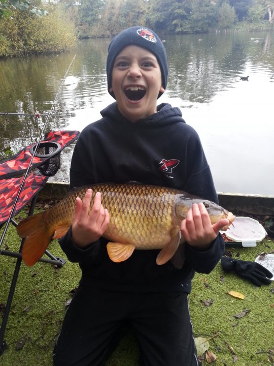 Starting fishing... - Page 2 - Sports - PistonHeads - In the image, an enthusiastic young boy is proudly displaying his catch, a large, tentative-looking fish. He's wearing a black hoodie adorned with a red star emblem. The boy is seated on a verdant floor, surrounded by tranquil wildlife and a vibrant red tent in the background. The overall setting exudes a sense of adventure and achievement, typical of a day spent fishing. The boy's joyous expression suggests a memorable experience in some serene, natural habitat.