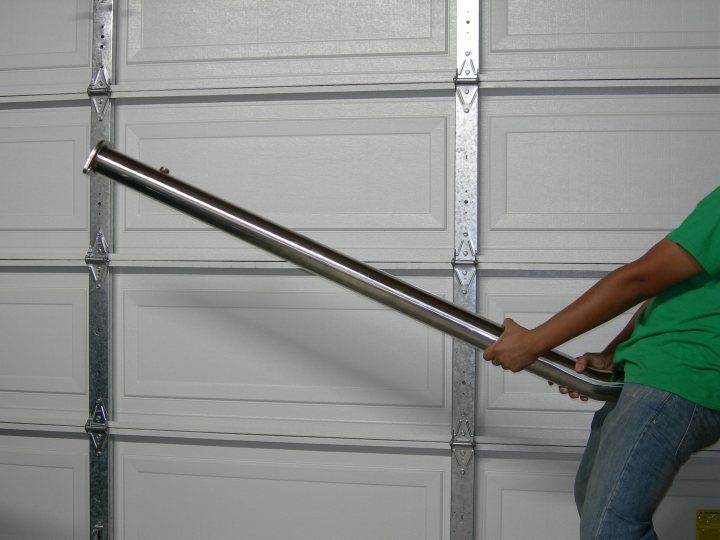 The image features a person in the process of pulling out a garage door. The person is dressed in a green shirt and is holding onto a long, metallic pole used to help open the door. The garage door itself has a white finish with multiple panels of opaque glass. The pole is resting against the lower part of the door, indicating an upward motion from the person. The style of the image is a freeze-frame photograph, capturing a precise moment of activity.