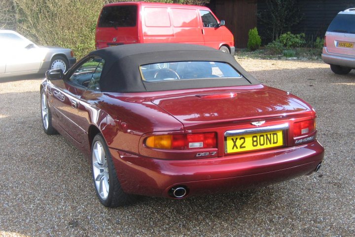 Pistonheads - The image depicts a red X2 Bond car parked on a gravel driveway. The car is emblazoned with a yellow license plate, indicating that it is ready to set out on its final mission. Adjacent to the car, there is a red van, suggesting a scene of organized endeavor or preparation. The setting appears to be an outdoor residential area, with foliage and other vehicles visible in the background. The image conveys a sense of adventure and excitement, characteristic of the iconic James Bond series.