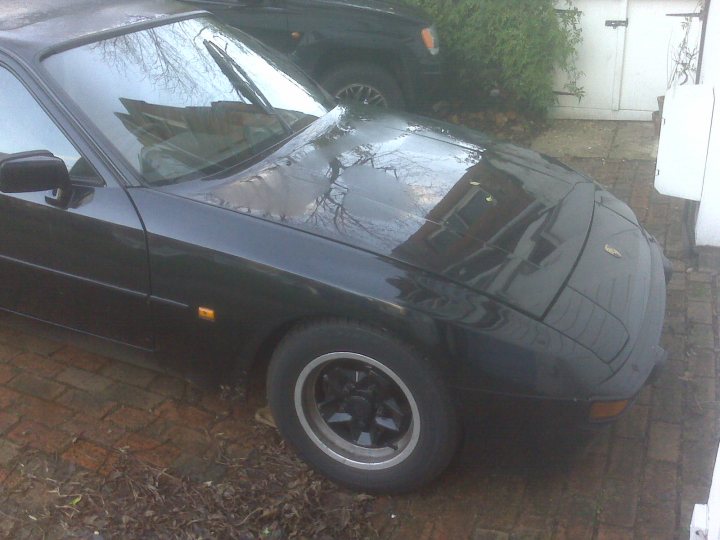 Pistonheads Hate Love - The image depicts a dark-colored sports car resting atop a driveway. The car's hood is slightly tucked in, while the headlight is clearly visible. The vehicle appears to be parked near a single brick wall, partially shaded. On the side of the driveway, some plants can be seen, adding a touch of greenery to the otherwise urban setting. In the backdrop, another vehicle can be glimpsed, further indicating that this is a residential area. The car's shiny exterior reflects the surrounding environment, hinting at the recent rainfall. Overall, the image captures a quiet, rainy day on a residential street.