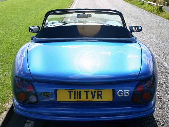 Wow! A sunny day... - Page 1 - North East - PistonHeads - The image displays a bright blue convertible car parked on a grassy area next to a road. The car has a vintage or retro styling, with a stripped-down roof and doors, hinting at a classic sports car design. The paint appears to be in a good condition, shining in the light. The license plate is visible, adding to the authenticity and individuality of the vehicle. The grass in the background suggests a non-urban outdoor setting, possibly during the day given the daylight visibility.