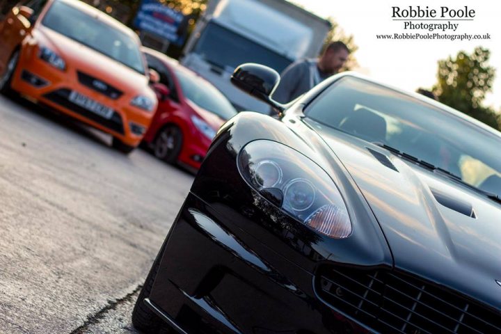 The GT Cafe - the ace cafe of the north - Aug meet - Page 1 - North West - PistonHeads - The image shows a collection of cars in what appears to be a parking lot at dusk or dawn, characterized by a soft, diffuse lighting. In the foreground, there is a striking black sports car with a prominent front design, suggesting a high-performance model. Among the other cars, there are a few compact vehicles and a large truck. No people are visible in the image, which focuses on the cars themselves and the ambient lighting from the setting or rising sun.