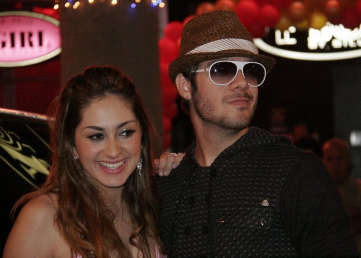 Fran - The image shows a young man and woman posing together. The man is wearing sunglasses and has a beard, while the woman has long hair and is smiling. They are indoors, possibly at a nightclub, as suggested by the dim lighting and the background featuring event-related decorations. The atmosphere seems to be festive and vibrant. Both individuals are facing the camera, engaging in what appears to be a happy moment.