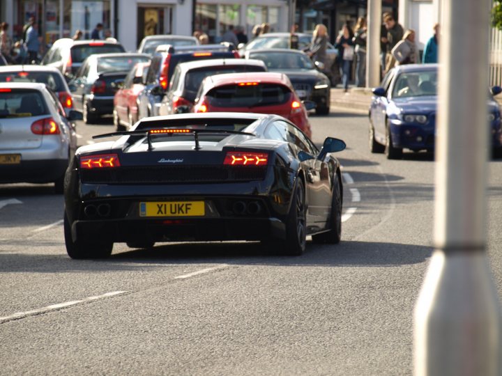 - Page 505 - North West - PistonHeads - This image captures a bustling city street. The main focus is a black sports car in the midst of heavy traffic. It's driving on the right side of the road, guided by its headlights and taillights. The sports car's license plate reads "X1 UKF". In the surrounding area, multiple other cars of various colors and models are visible, mirroring the busy nature of the road. Pedestrians can also be seen navigating the sidewalks on either side of the street, adding to the dynamic urban scene. To the right of the frame, a silver pole stands tall against the cityscape.