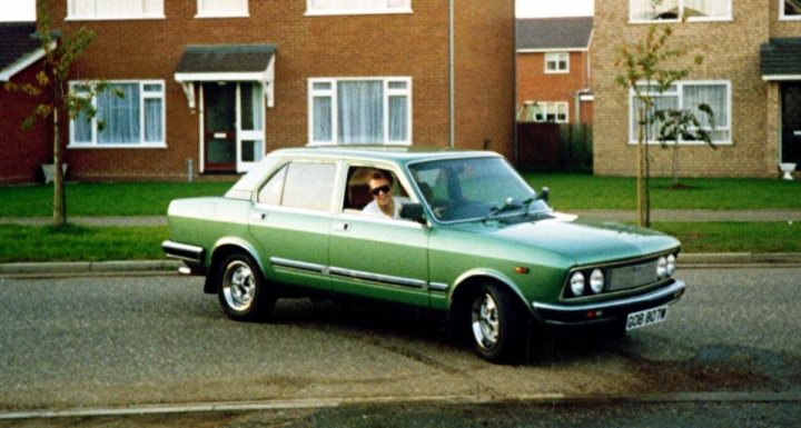 1977 Fiat 132  - Page 1 - Readers' Cars - PistonHeads - In the image, a British Ford Escort MK1 is the focal point, captured in a stationary position on the side of a road. The car's vibrant green body contrasts with the plain suburban street it's parked on. Adjacent to the car is a house, identifiable by a small garden and a tree planted right in front of it. A single person can be seen walking beside the car, adding a sense of scale to the scene. In the sky above, the sun is setting, casting a warm glow over the entire image, enveloping the scene in a soft, golden light.