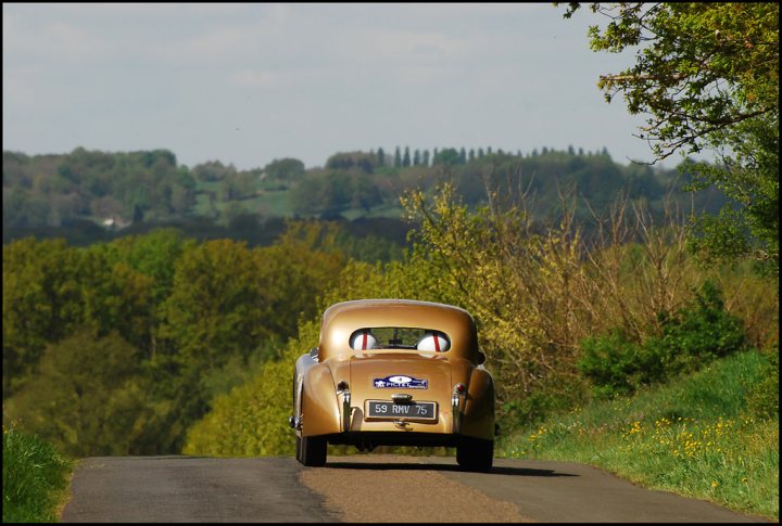 The Tour Auto came to town - Page 1 - Classic Cars and Yesterday's Heroes - PistonHeads - The image depicts a serene countryside scene. Dominating the foreground is a classic, vintage car that has an old-world charm. The car is parked on the side of a paved road, possibly indicating a stop or pause. The car's position near the edge of the road gives an impression of it just having completed the journey or is preparing to embark on one. The backdrop features an expanse of a wooded area with a hill in the distance, adding a natural and tranquil touch to the overall scene.