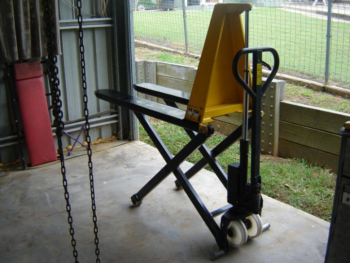 Best way to lift a Radical high enough to work on - Page 1 - Radical - PistonHeads - The image features a large yellow industrial lift with a platform and two vertical supports. The lift is situated on a concrete ground near the entrance of a structure with a chain-link fence in the background. There is a bench, also in yellow, in the foreground with a backrest, inviting the viewer to sit. The setting is outdoors, and the overall scene gives off an industrial or maintenance vibe.