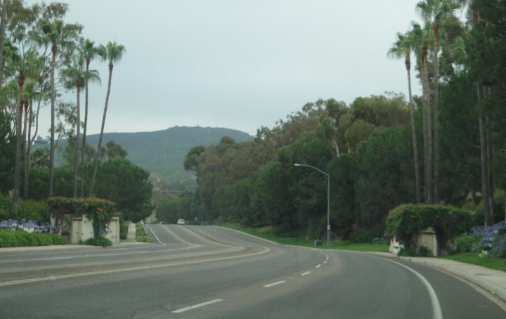 The image presents an empty road that extends into the distance, bordered by verdant trees on both sides. Above, the sky is overcast, suggesting a cloudy day. The trees on the right side of the road are notably taller and more densely packed, creating a natural canopy over the road. In the distance, the faint outline of a mountain can be seen. The road itself appears to be well-maintained, with clear lines marking the lanes, and there is no visible traffic or pedestrians at present.