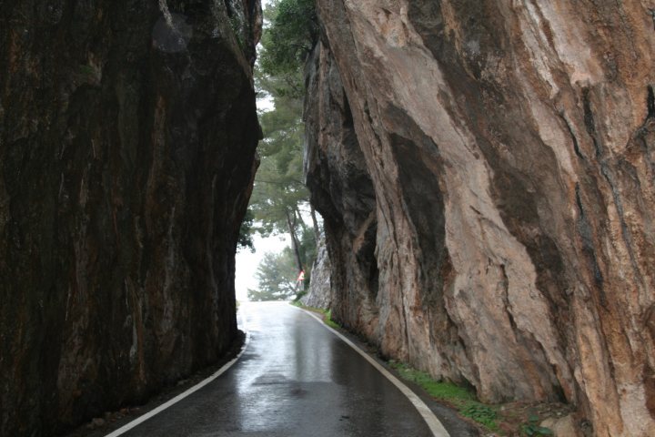 VII Rally Clasico Isla Mallorca. - Page 1 - General Gassing - PistonHeads - The image depicts a narrow, tree-lined road that winds through the sides of imposing cliffs. The road is wet and appears to be reflective, adding to the sense of isolation and mystery. The cliffs rise steeply on either side, with trees clinging to the narrow passage between them. The scene is dramatic, with the road receding into the distance, offering glimpses of the sky and trees beyond the cliffs. There is a slight mist or fog that gives the scene an ethereal quality.