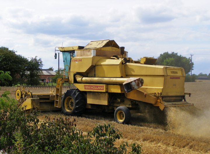 RE: Mr Big Stuff: Claas Lexion 530 Combine - Page 3 - General Gassing - PistonHeads - The image depicts a large yellow agricultural harvester or combine working in a field of crops. The machine is covered in dust, indicating it's actively engaged in harvesting. In the field, a group of green bushes is visible, adding texture to the agricultural scene. Also, a large building with a blue roof starkly contrasts the natural environment, hinting at human presence or infrastructure nearby. The combine, with its yellow body and black tires, stands out prominently against the greenery and dirt. The sky above is filled with clouds, suggesting a cloudy day.